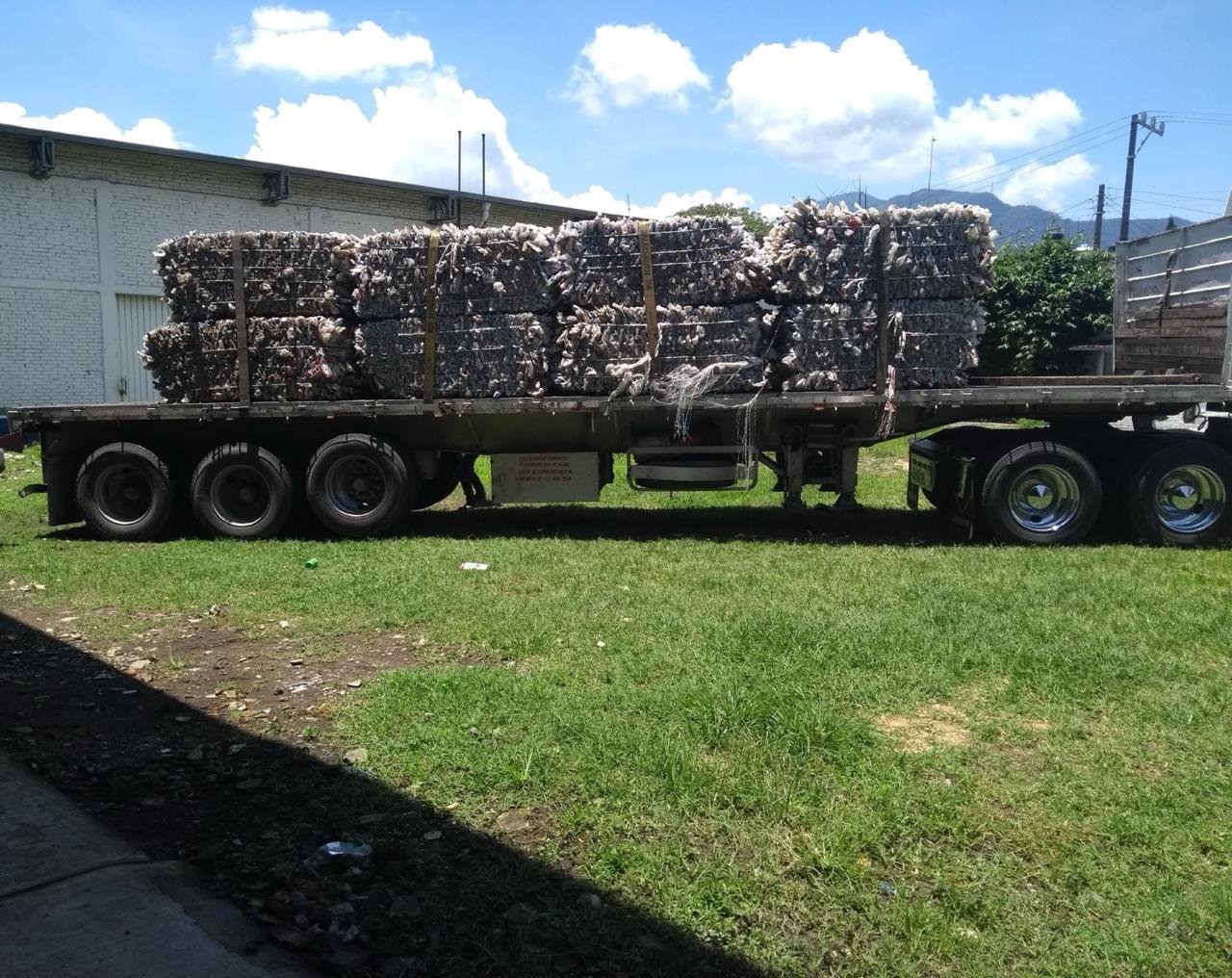 Large flatbed truck loaded with stacked bales of recycled materials on grass with mountains and white building in background