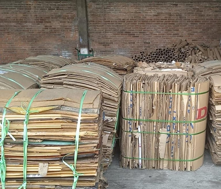 Stacks of bundled cardboard and paper waste tied with green straps against a brick wall, ready for recycling